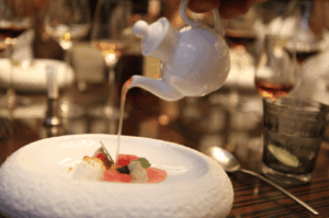 A server pours aromatic broth from a white teapot onto a meticulously plated dish featuring radish slices, mousse dollops, and edible flowers, set on a textured white plate in a refined dining setting. The elegant tablescape—complete with wine glasses and lime-garnished water—evokes the theatrical precision of Michelin dining, contrasting with the broader ambiance of fine dining