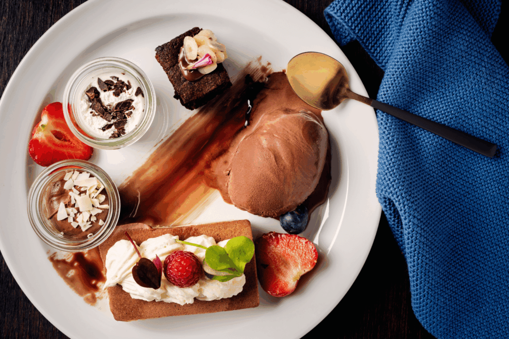 Artful Singapore dessert medley plated on white porcelain—featuring chocolate ice cream, layered parfaits, brownie with floral garnish, and a raspberry-topped pastry—framed by fresh berries, a gold spoon, and a blue napkin, embodying the elegance, precision, and sensory delight of a signature fine dining finale.