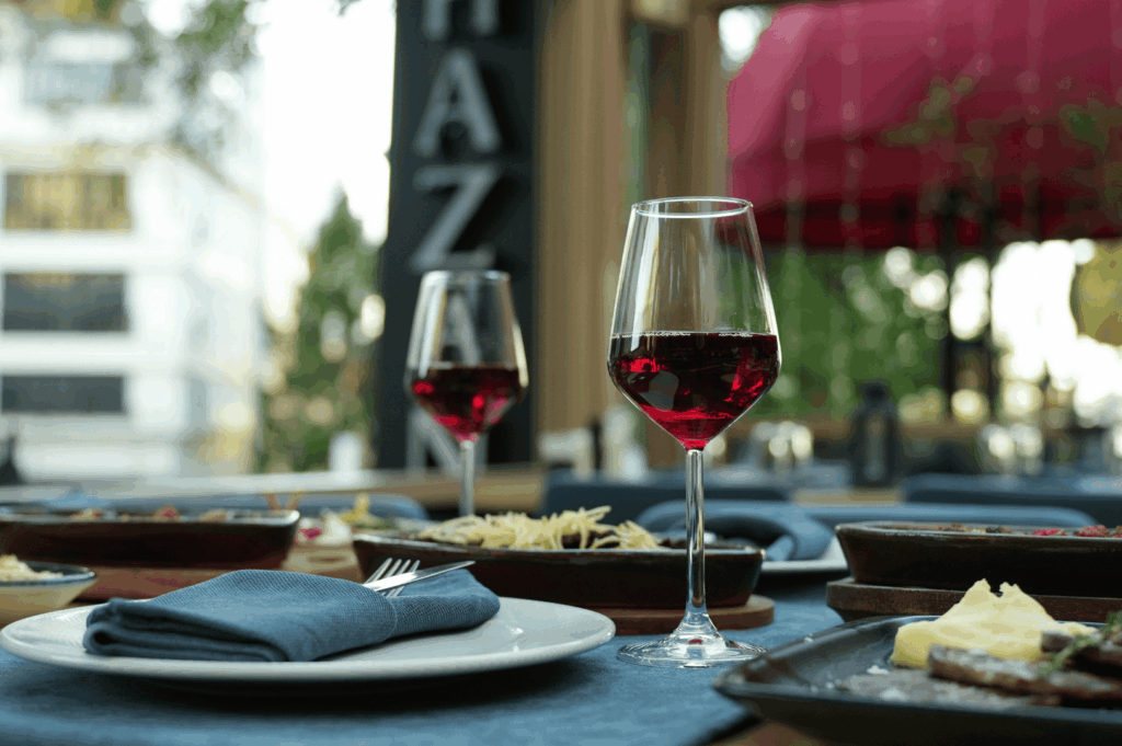 Open-air Singapore fine dining table dressed in blue linen, featuring pasta, cheeses, and red wine under a red canopy with lush greenery—evoking the relaxed elegance, sensory richness, and alfresco charm that define a signature culinary experience in Southeast Asia’s vibrant hospitality scene.