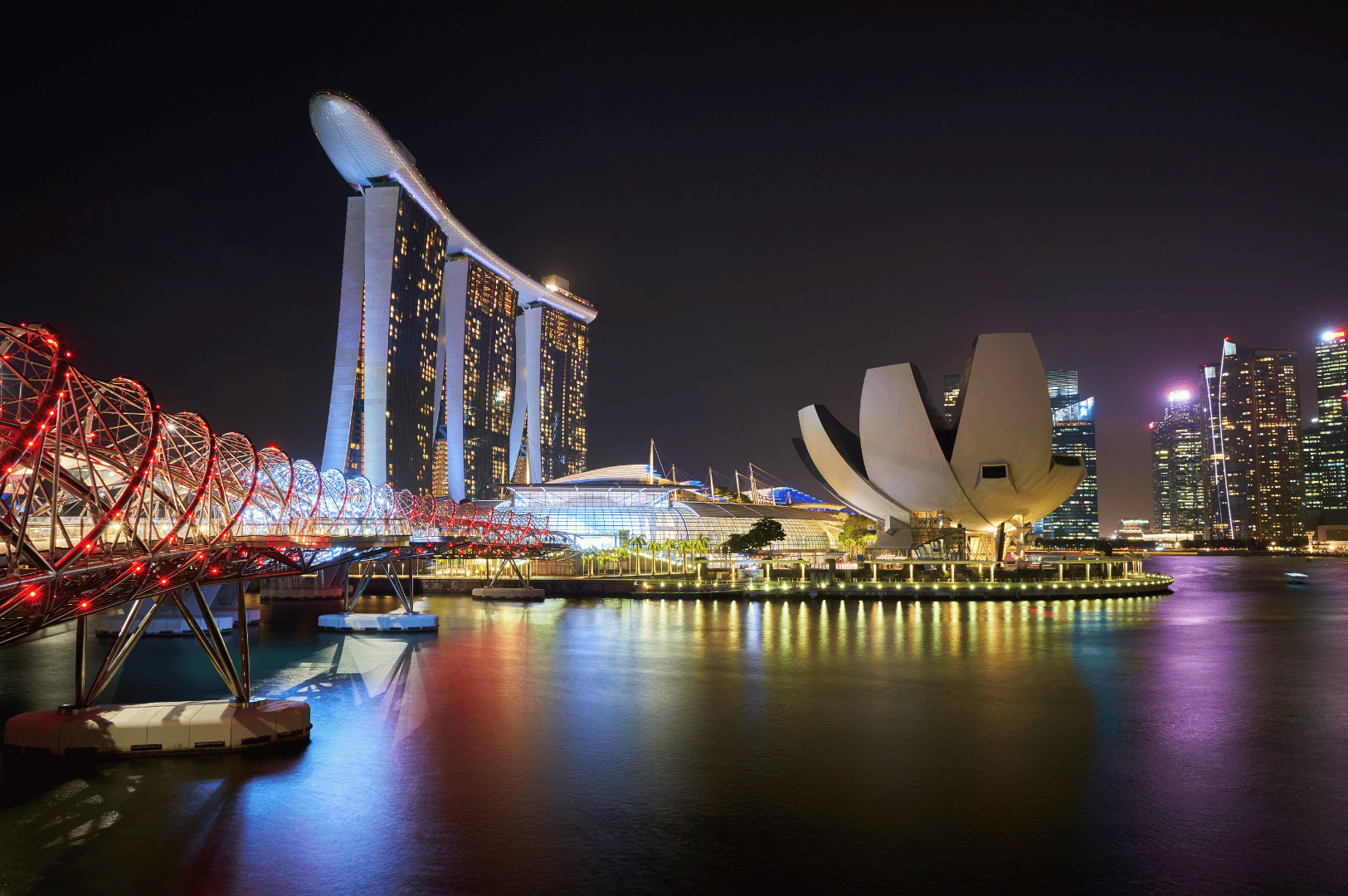 Nighttime view of Marina Bay in Singapore, featuring the illuminated Marina Bay Sands hotel, ArtScience Museum, and Helix Bridge reflected in the bay’s waters—an iconic urban skyline that frames some of the city’s most scenic waterfront restaurants.