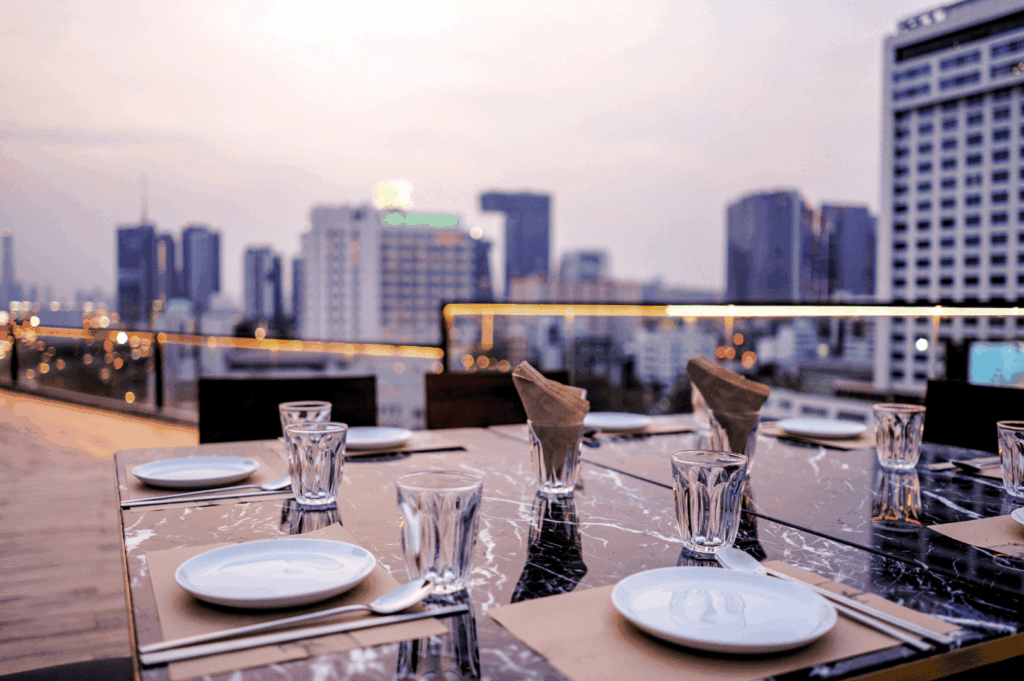 Elegant rooftop dining setup overlooking Singapore’s city skyline at dusk, with marble table settings and warm urban glow—capturing the refined ambiance of high-altitude restaurants that pair panoramic views with sophisticated cuisine.