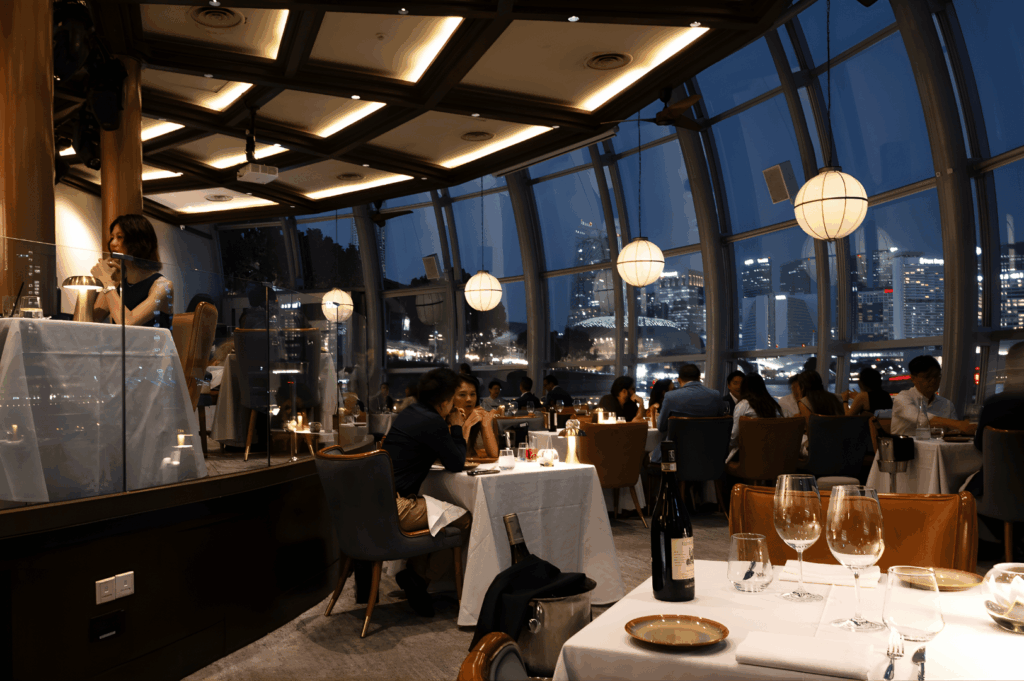 Upscale restaurant interior with floor-to-ceiling windows framing Singapore’s glittering skyline at night; warm lighting, elegant furnishings, and candlelit tables evoke a luxurious dining experience where city views meet refined hospitality.