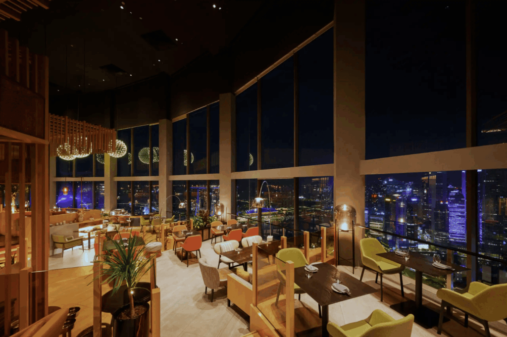 Chic high-rise restaurant lounge with panoramic nighttime views of Singapore’s cityscape; warm pendant lighting, plush seating, and indoor greenery create a cozy yet refined atmosphere—ideal for savoring skyline dining in style.