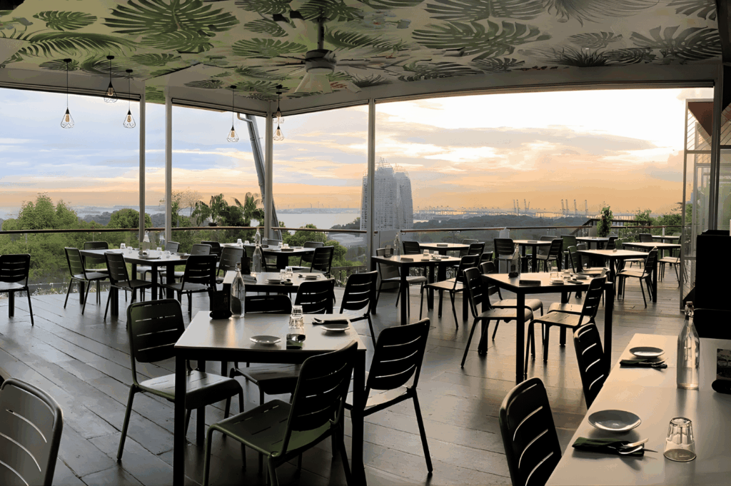 Open-air restaurant with leafy ceiling accents and panoramic harbor views at sunset; black tables, minimalist place settings, and warm hanging lights evoke a relaxed yet refined dining experience where nature and cityscape meet.