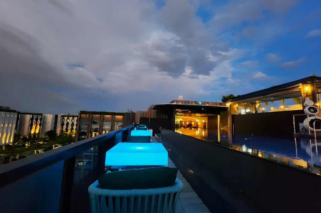 Rooftop view at dusk with glowing blue tables leading to a modern bar. A cloudy sky adds a serene, relaxed atmosphere. Urban buildings in the background.