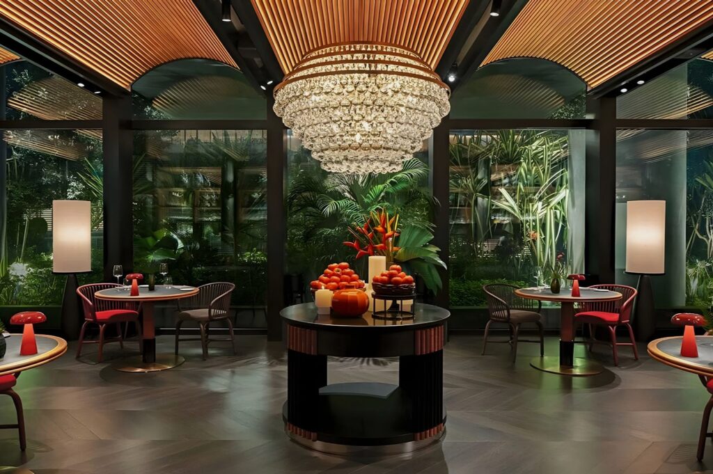 The upscale dining area at Quenino, Singapore, presents ambient illumination, glass-enclosed greenery, circular tables, red seating, and a prominent fruit centerpiece as focal decor.