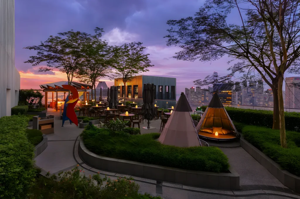 Rooftop terrace at sunset, featuring modern seating, teepee tents, and ambient lighting. Surrounded by greenery and city skyscrapers in the background.