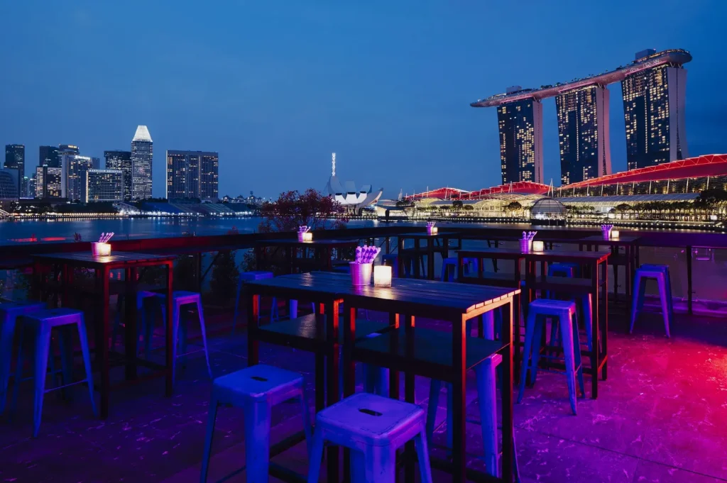 Rooftop bar at dusk with tables and stools under purple lighting, overlooking a city skyline and iconic building. A calm, vibrant atmosphere.