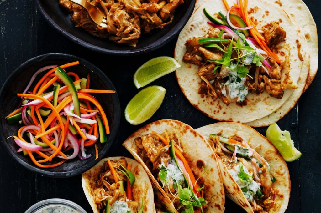 Close-up of Rendang Tacos topped with herbs and onions, served with lime wedges and a bright carrot-cucumber slaw.