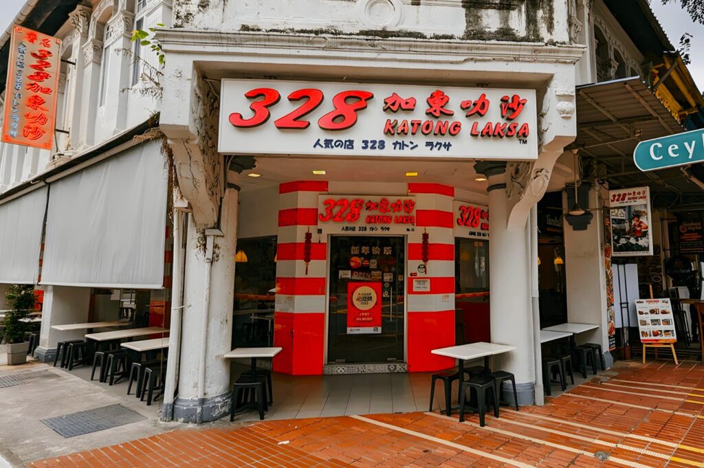 Street view of 328 Katong Laksa, a brightly colored restaurant with red and white signage. Outdoor seating with black stools creates a vibrant, inviting atmosphere.