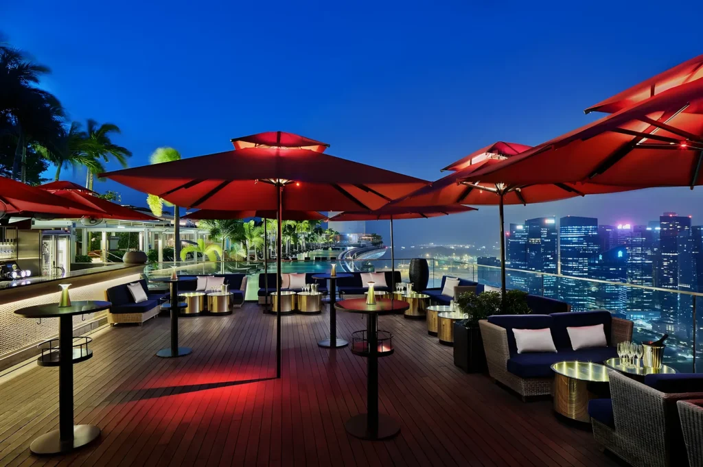 Rooftop lounge at night with red umbrellas, wicker seating, and city skyline view. Ambient lighting creates a relaxing, upscale atmosphere.