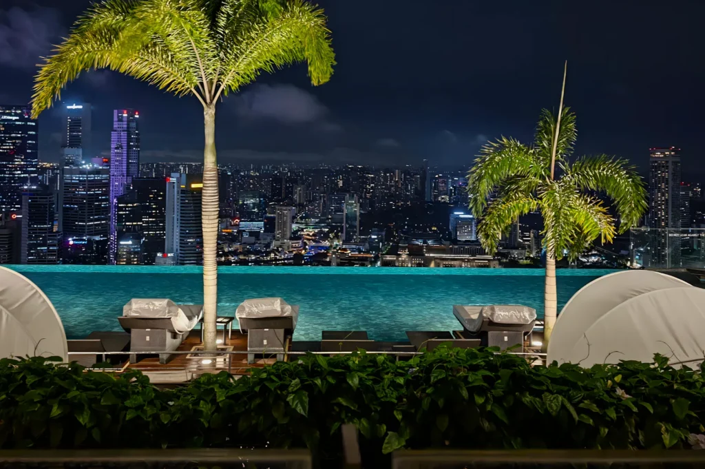 Rooftop pool overlooks a vibrant city skyline at night. Palm trees frame the view, and lounge chairs line the poolside, creating a luxurious ambiance.