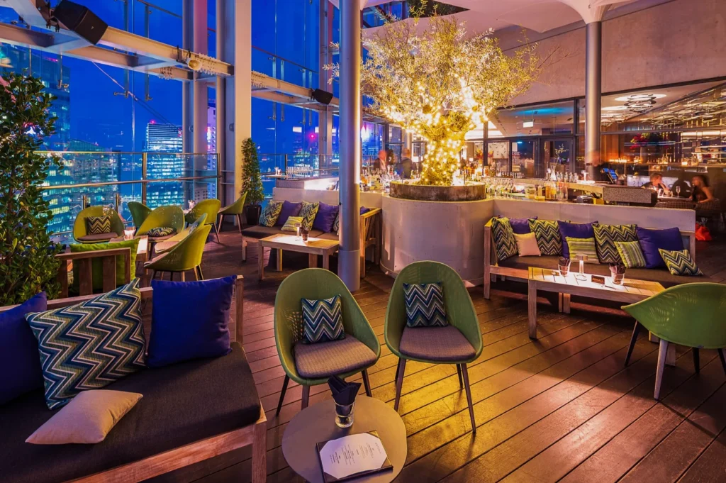 Chic rooftop lounge at night with glowing tree centerpiece, plush seating with colorful pillows, and cityscape view through large windows. Cozy ambiance.