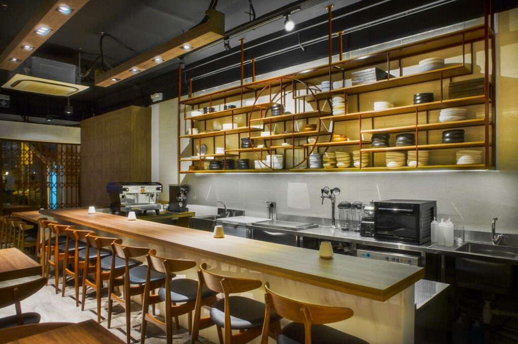 A sleek open-kitchen dining area at Restaurant Ibid, Singapore, with a long wooden bar, a row of wooden stools, and a wall of neatly arranged plates.