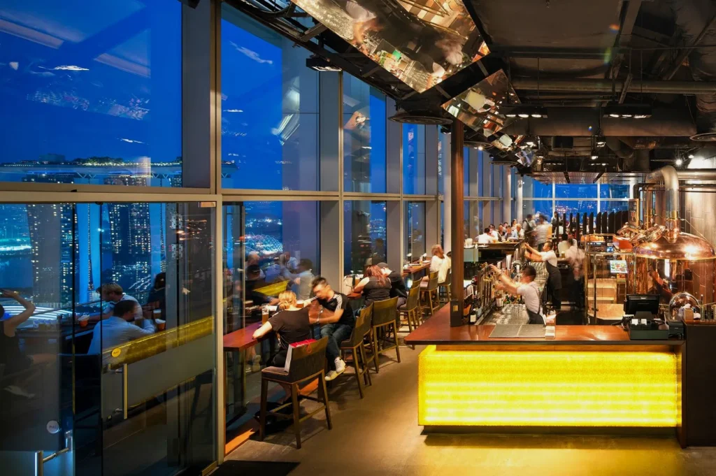 Modern rooftop bar with a glowing counter, featuring patrons seated at tables enjoying drinks. Large windows offer a stunning city skyline view at dusk.