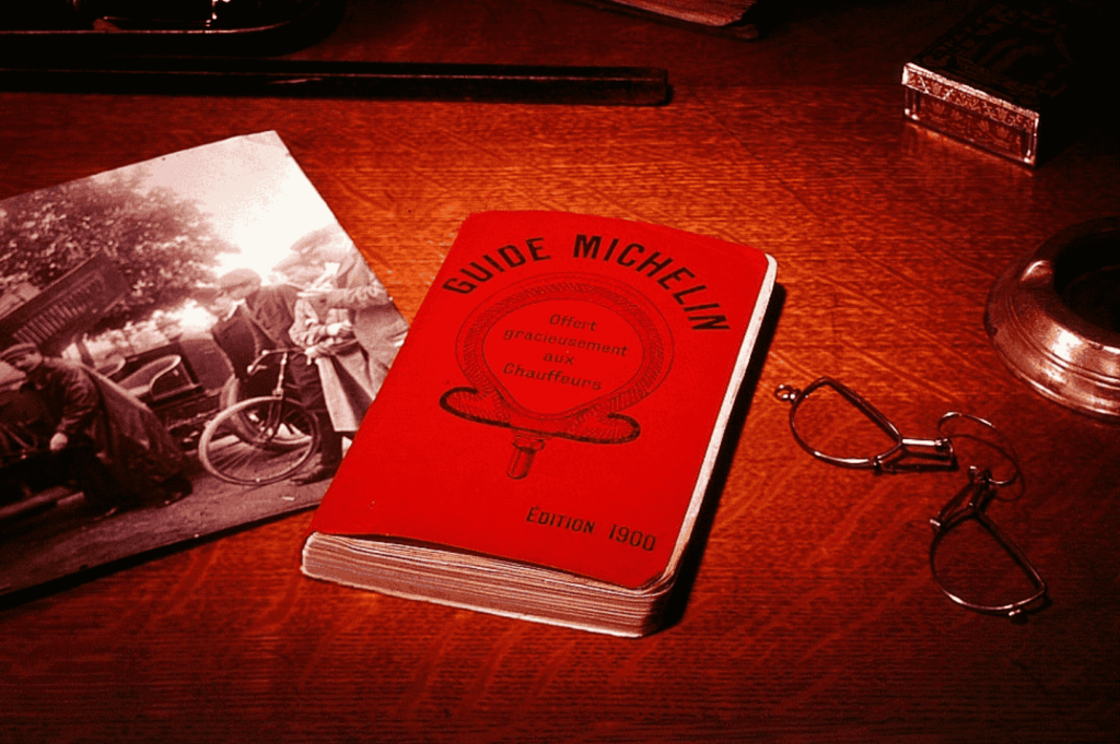 A vintage 1900 edition of the Michelin Guide displayed on a wooden desk beside round eyeglasses, an inkwell, and a black-and-white photo of early automobile diners—capturing the origins of the michelin star restaurant guide and the evolution of michelin star restaurants. This historical artifact marks the beginning of the culinary journey toward fine dining excellence, where michelin inspectors developed the five criteria that now define one michelin star, two michelin stars, and three michelin stars. From the first michelin star awarded to a fine dining restaurant to today’s three star restaurant experiences at Summer Pavilion, Saint Pierre, and Marina Bay, the michelin guide has shaped the hospitality industry and inspired world renowned chefs like chef louis han, chef yew eng tong, head chef takuya yamashita, chef owner emmanuel stroobant, chef denis lucchi, and chef christophe lerouy. Whether crafting a tasting menu with sea urchin and hairy crab, or a lunch menu featuring nigiri sushi and premium cuts, these fine dining establishments offer diners exceptional cuisine, seasonal ingredients, and innovative dishes. Michelin introduced the michelin green star and bib gourmand to highlight sustainability and good restaurant practices, while young chefs and sous chefs continue to pursue culinary excellence and express the chef’s personality through cooking techniques and global influences. This image reflects the special journey from good restaurant to culinary haven, where all the restaurants strive for gastronomic excellence and the coveted first star.