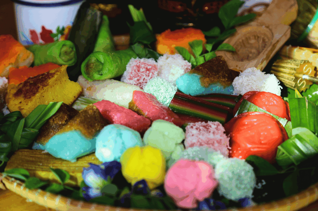 Assorted traditional kuih arranged on a woven tray lined with banana leaves—vibrant pink, green, blue, and yellow bite-sized sweets made from glutinous rice, coconut milk, and pandan, some dusted with shredded coconut. This colorful medley reflects the artisanal roots and evolving aesthetics of Peranakan dessert culture in Singapore’s fine dining scene.