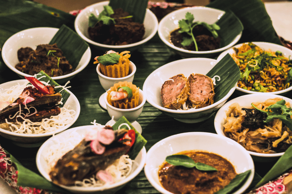 Traditional Southeast Asian tasting tray featuring an array of small white bowls filled with richly spiced meats, vegetables, noodles, and sauces—each garnished with herbs, chilies, or crispy toppings. Presented on banana leaves atop a circular platter, this communal spread reflects the layered textures and bold flavors that inspire modern Peranakan fine dining in Singapore.