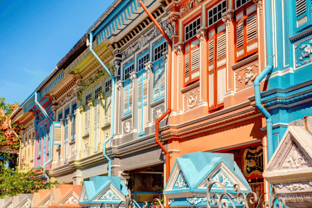 Row of pastel-hued Peranakan shophouses in Singapore, featuring ornate window carvings, shuttered facades, and vibrant architectural details in blue, yellow, green, and red. The decorative fence in the foreground adds charm, while the preserved heritage buildings evoke the cultural roots that shape the aesthetics and storytelling of modern Peranakan fine dining.