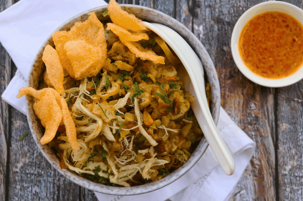 Indonesian-style chicken porridge (bubur ayam) served in a ceramic bowl on rustic wood—topped with shredded chicken, fried shallots, herbs, and crispy crackers, with sambal on the side. This comforting dish bridges Southeast Asian street food traditions with the refined textures and plating sensibilities shaping Singapore’s modern Peranakan fine dining
