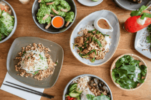 A vibrant spread of Vietnamese-inspired fusion dishes arranged on a rustic wooden table, featuring grilled meats, vermicelli noodles, fresh herbs, and dipping sauces. The composition blends Southeast Asian street food aesthetics with modern plating, highlighting Singapore’s culinary harmony between tradition and innovation. A red citrus drink garnished with mint and lime adds a refreshing contrast, evoking the sensory richness of East-meets-West dining culture.