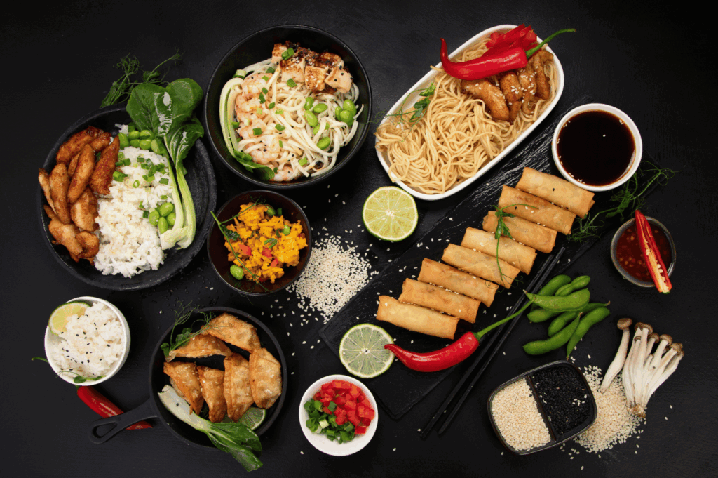 A visually striking assortment of Asian dishes arranged on a black surface, featuring rice bowls with grilled chicken, noodle soups with shrimp, spring rolls, dumplings, curry, and vibrant garnishes like chili peppers, lime slices, and enoki mushrooms. The interplay of colors, textures, and plating reflects Singapore’s fusion philosophy—where culinary traditions from across Asia are reimagined with precision, creativity, and bold flavor harmony.