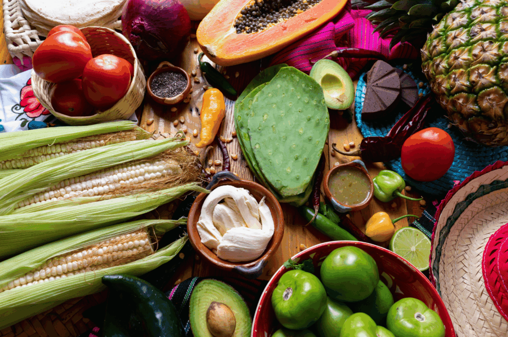 A vibrant tableau of traditional Mexican ingredients—corn, tomatoes, avocados, chili peppers, mole, and tropical fruits—artfully arranged on a rustic wooden surface. The composition celebrates culinary heritage and visual abundance, echoing Singapore’s fusion ethos where global flavors meet local innovation. Textiles and sombreros add cultural texture, inviting a sensory dialogue between Latin American tradition and Southeast Asian reinterpretation.