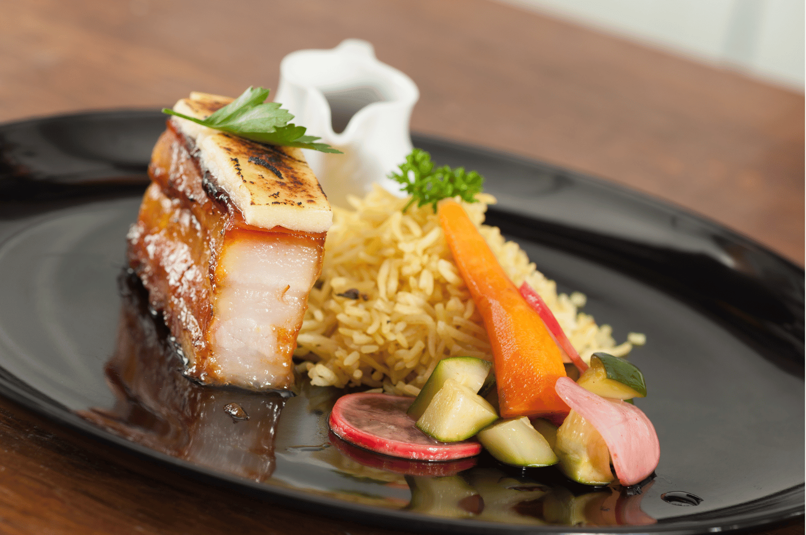 An elegantly plated fusion dish featuring caramelized pork belly crowned with grilled cheese and parsley, paired with seasoned rice and a vibrant medley of pickled vegetables. Set against a matte black plate, the composition evokes fine dining sophistication while nodding to Southeast Asian comfort food. A white sauce container in the background hints at layered flavor, embodying Singapore’s culinary dialogue between heritage and innovation.