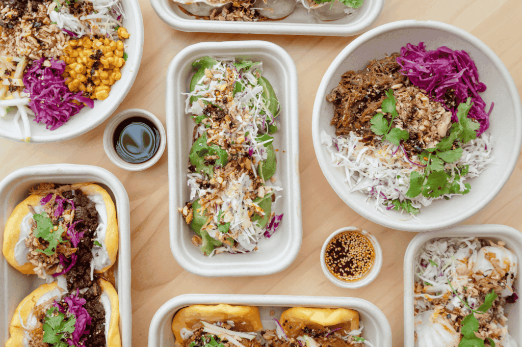 A dynamic spread of fusion street food featuring tacos, bao buns, and salad bowls, layered with vibrant ingredients like purple cabbage, grilled corn, pulled meats, and sesame-topped sauces. The mix of Latin American and Asian elements—crispy onions, cilantro, soy-based dips—embodies Singapore’s culinary crossroads, where global flavors are reimagined with local flair. The light wooden table and artful plating evoke casual sophistication and multicultural abundance.