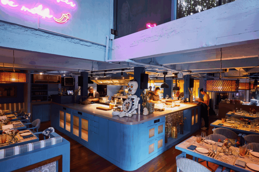 A stylish open-kitchen restaurant interior featuring a vibrant blue island where chefs prepare food in full view of diners. Warm ambient lighting, perforated hanging lamps, and eclectic decor—including a playful skeleton figure—create a lively, contemporary atmosphere. Neon signage and upholstered seating evoke Singapore’s fusion dining culture, where global design sensibilities meet local hospitality traditions in a space that celebrates culinary creativity.