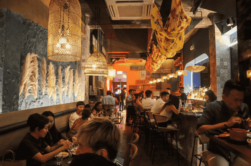 A lively, design-forward restaurant interior featuring woven pendant lamps, pampas grass wall accents, and colorful ceiling drapes. Patrons gather around tables and a well-stocked bar, creating a warm, social atmosphere that blends bohemian charm with urban sophistication. The eclectic decor and vibrant energy reflect Singapore’s fusion ethos—where global aesthetics and local hospitality converge in immersive dining experiences.