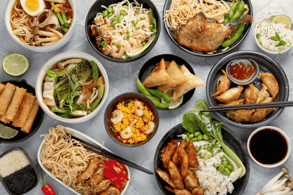 A vibrant array of Asian-inspired dishes arranged on a communal table, featuring noodle bowls with grilled duck and boiled eggs, spring rolls with lime, dumplings, fried rice, and grilled chicken with bok choy and edamame. The composition celebrates the diversity of textures and flavors—crispy, savory, umami-rich—reflecting Singapore’s fusion philosophy where regional traditions converge in bold, modern expressions of comfort food.