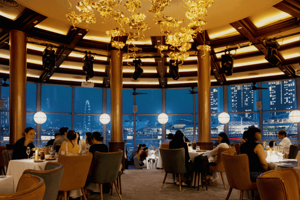 Elegant evening dining scene in a high-rise Singapore restaurant with golden floral chandeliers, plush seating, and panoramic windows framing the illuminated waterfront skyline; a lively yet refined ambiance blending architectural beauty with upscale urban hospitality.