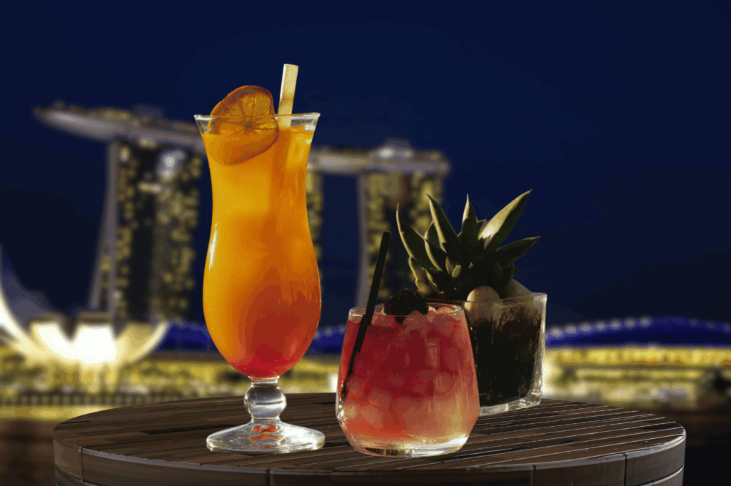 Three colorful cocktails on a wooden surface with Singapore’s Marina Bay Sands illuminated in the nighttime background; vibrant orange, pink, and green drinks with tropical garnishes evoke a festive, luxurious mood against the city’s shimmering skyline.