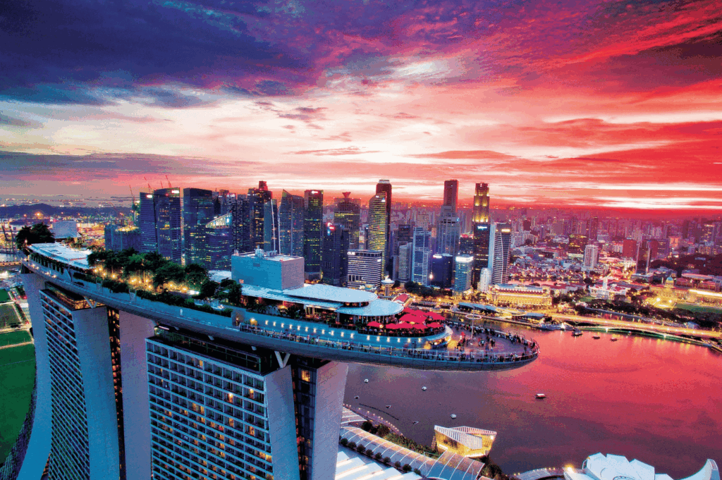 Aerial sunset view of Marina Bay Sands in Singapore, with its iconic sky park aglow in red lights and bustling with visitors; dramatic hues of purple, orange, and blue reflect across calm bay waters, framing the city’s modern skyline in a breathtaking blend of architecture and atmosphere.