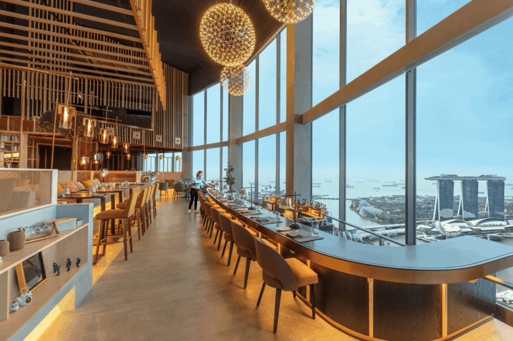 Elegant high-rise restaurant interior with panoramic floor-to-ceiling windows framing Singapore’s Marina Bay Sands and coastal skyline; warm lighting, sculptural chandeliers, and plush seating evoke refined ambiance and elevated dining with a view.