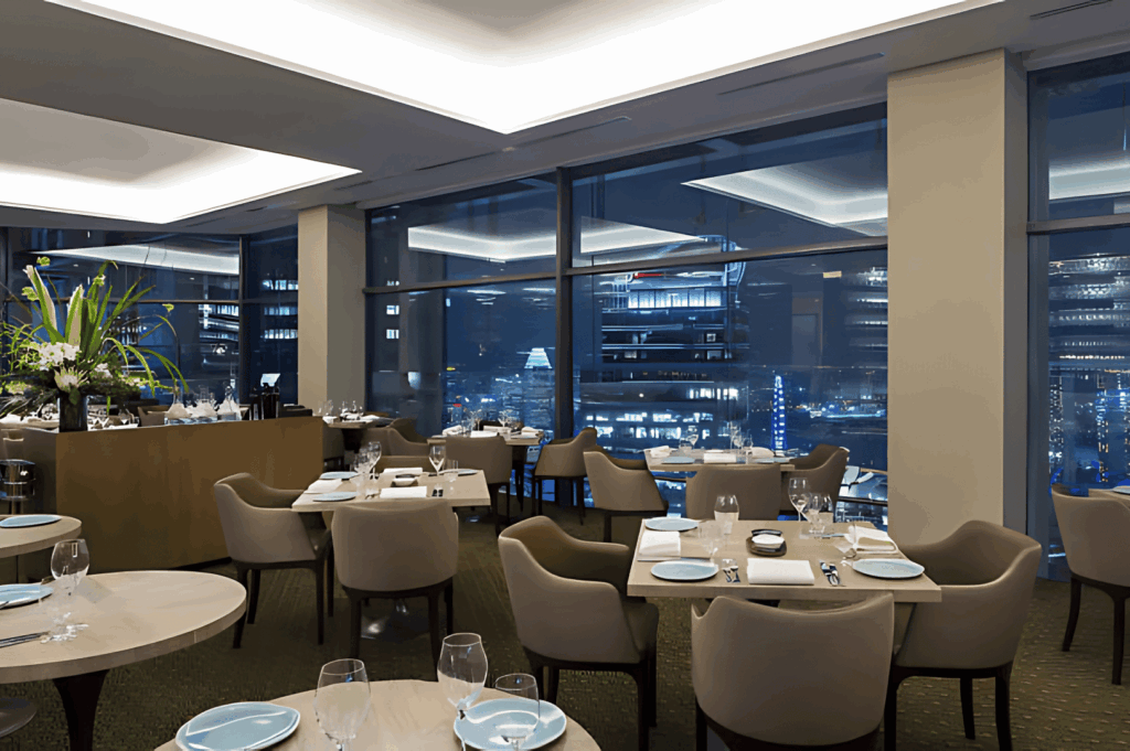 Upscale restaurant interior with floor-to-ceiling windows revealing Singapore’s glittering nighttime skyline; soft ambient lighting, elegant table settings, and floral accents create a refined, intimate atmosphere for elevated city-view dining.