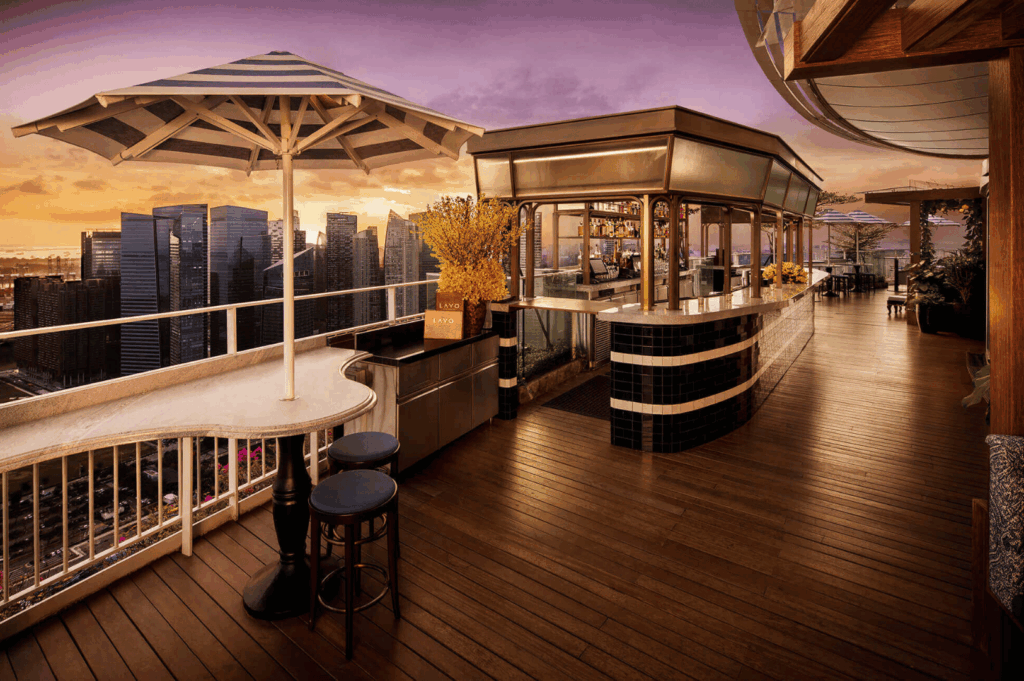 Stylish rooftop bar in Singapore at sunset, featuring sleek counters, high stools, and yellow floral accents under wide umbrellas; warm purple-orange sky and towering cityscape create a vibrant yet relaxed setting for elevated urban dining.