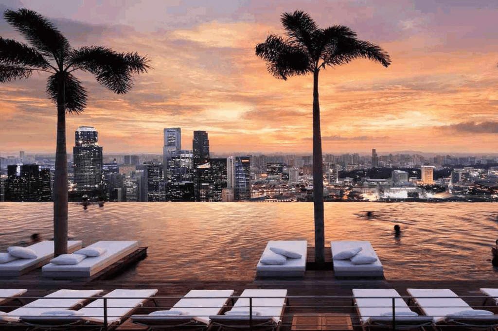 Sunset view from a rooftop infinity pool in Singapore, where warm hues of orange and pink blend with city skyline silhouettes; cushioned lounge chairs and swaying palm trees frame a tranquil scene of luxury, leisure, and elevated urban escape.
