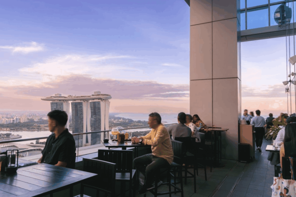 Lively rooftop restaurant in Singapore during sunset, with guests dining against a panoramic skyline featuring Marina Bay Sands; soft blue-pink sky and elevated city views create a vibrant, social atmosphere blending luxury, architecture, and urban leisure.