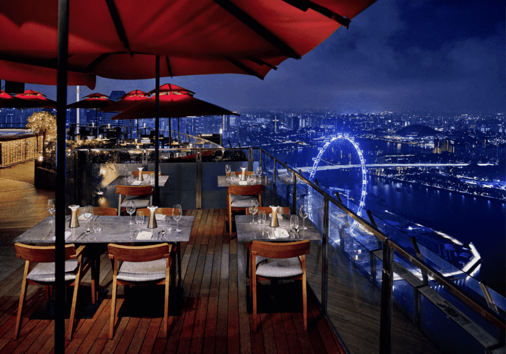 Nighttime rooftop dining with a view in Singapore featuring alfresco balconies, candlelit tables, and signature cocktails under red umbrellas, overlooking the city skyline and panoramic views of Marina Bay. This romantic restaurant offers stunning views of Marina Bay Sands, Marina Bay Financial Centre, and the illuminated city lights of Downtown Singapore. Among the best rooftop restaurants and rooftop bars in the Marina Bay area, near Sentosa Island, Mount Faber Peak, Ocean Financial Centre, Customs House, Collyer Quay, Civic District, Raffles Place, and OUE Bayfront. Ideal for happy hour, special dinner, weekday lunch, or private dining rooms, with a menu of contemporary Asian cuisine, modern Asian cuisine, European classics, small plates, freshest seafood including Maine lobster, and dishes curated by executive chef Sam Chin and executive chef Oliver Hyde. A must-visit destination for rooftop dining and dining with a view Singapore, featuring an impressive selection of food and drinks in a luxurious dining room setting.