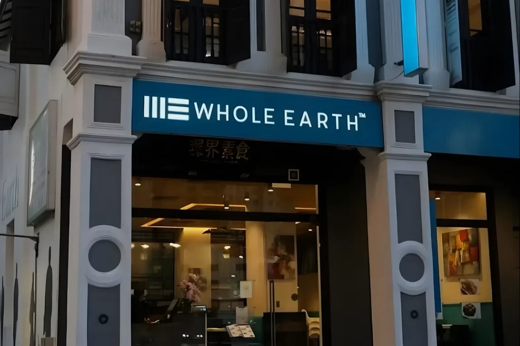 Storefront of "Whole Earth" restaurant with blue signage, framed by white columns. Warm interior lighting visible through large glass doors. Calm atmosphere.