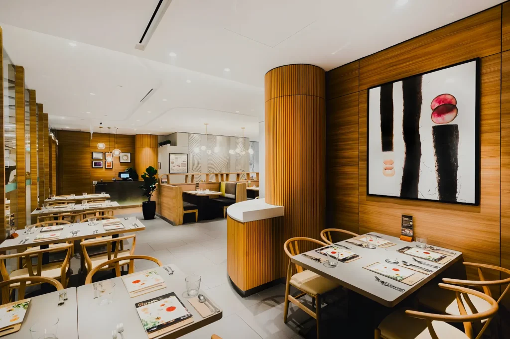 Modern restaurant interior with wooden accents, soft lighting, and neatly set tables. Art adorns the walls, creating a warm, elegant atmosphere.