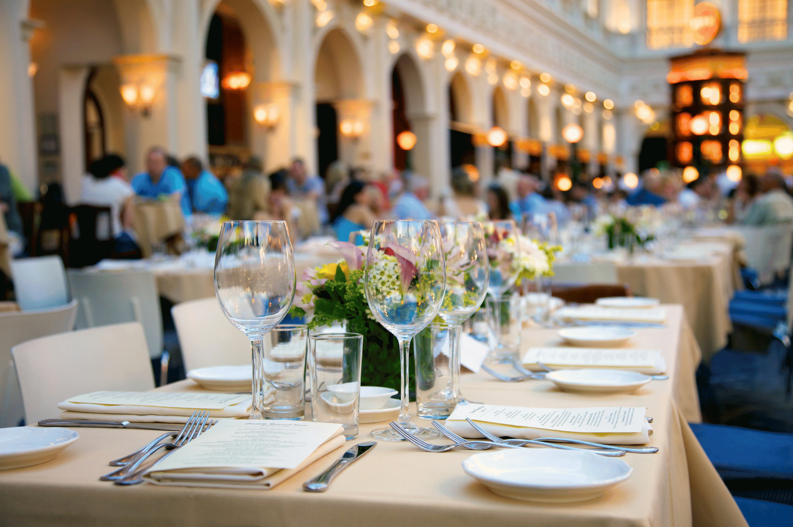 Elegant outdoor dining setup at a luxury Singapore venue, featuring beige tablecloths, floral centerpieces, and meticulously arranged tasting menus. Guests seated in the background enjoy a curated multi-course experience under warm architectural lighting, evoking the sophistication and sensory richness of Singapore’s best tasting menus.