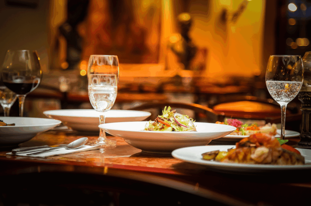 Polished wooden table set for fine dining in a warm, art-filled Singapore restaurant. Plates of gourmet dishes—leafy salad, grilled meat with vegetables—are paired with red wine, sparkling water, and elegant glassware. The softly lit background, featuring blurred sculptures and artwork, enhances the intimate, curated atmosphere of a tasting menu experience.