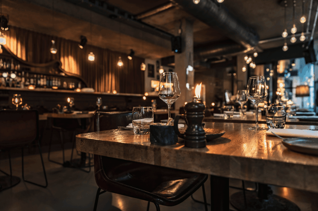 Dimly lit interior of an upscale Singapore restaurant with modern industrial design—exposed pipes, concrete walls, and warm hanging bulbs. Wooden tables are elegantly set with wine glasses, cloth napkins, and a glowing candle, creating a cozy, romantic atmosphere that invites reflection at the end of a curated tasting journey.