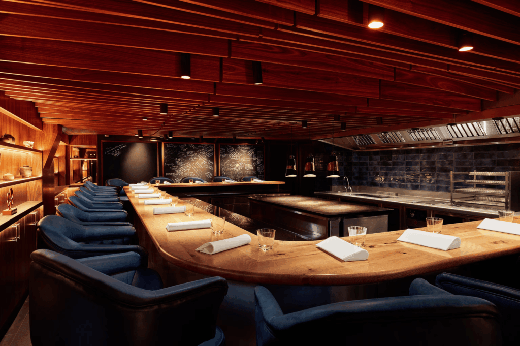 An immersive chef’s table setting in a high-end restaurant, featuring a curved wooden counter lined with plush blue chairs, each place set with a white napkin and glassware. The open kitchen behind the counter showcases stainless steel appliances, a grill, and a dark brick backsplash, inviting guests into the heart of the culinary process. Warm lighting from recessed fixtures and wooden ceiling slats enhances the intimate ambiance, while curated shelves with bottles and decor add a touch of sophistication. The scene captures the transformative power of dining where the boundary between kitchen and guest dissolves, offering a front-row seat to culinary artistry.