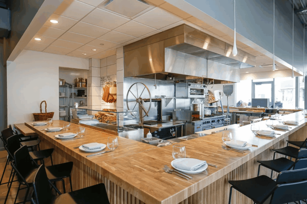 A bright, modern chef’s counter in an open-concept restaurant kitchen, featuring light wood surfaces and black chairs arranged for intimate dining. Each place is set with white plates, wine glasses, and silverware, ready for guests to engage with the culinary process. Behind the counter, stainless steel appliances, a rotisserie with skewered meats, and neatly organized shelves of ingredients evoke a sense of precision and craft. Natural light floods the space through large windows, creating a clean, inviting atmosphere where the boundary between kitchen and table disappears, immersing diners in the rhythm of preparation and flavor.
