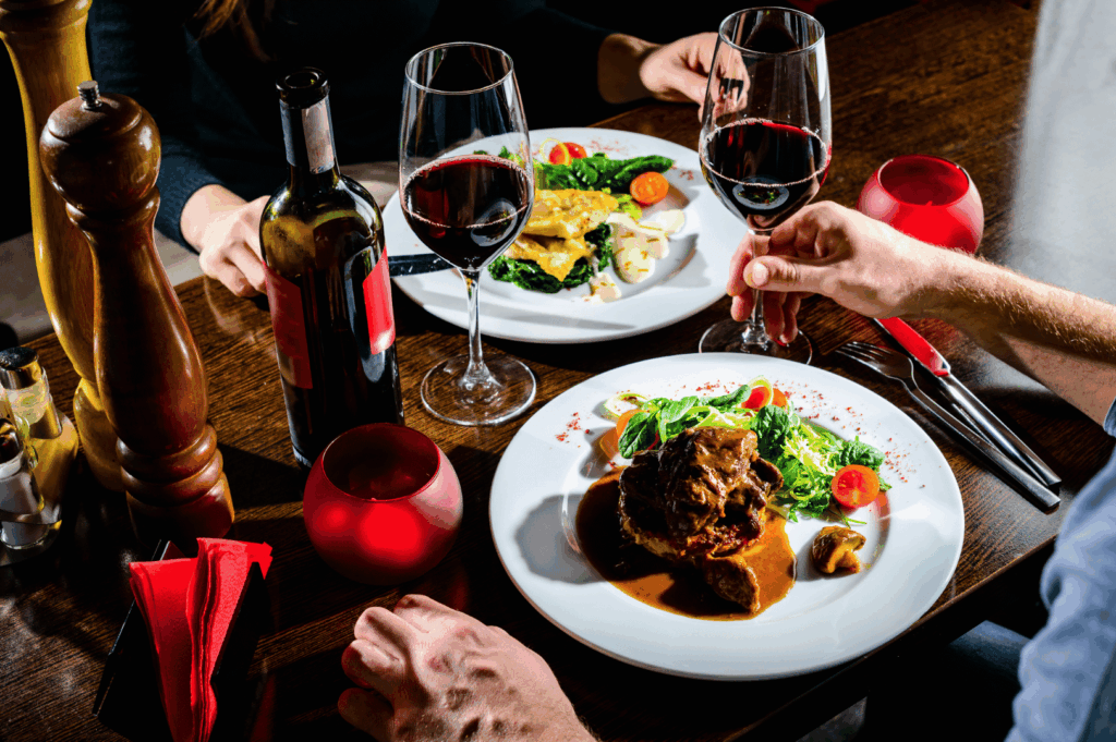 Warm restaurant scene with two diners toasting red wine over plates of meat, fish, mashed potatoes, and greens. Surrounded by candlelight, wine bottles, and red napkins, the image captures the celebratory intimacy and shared joy of fine dining—contrasting Michelin’s structured prestige with the emotional richness of personal connection.