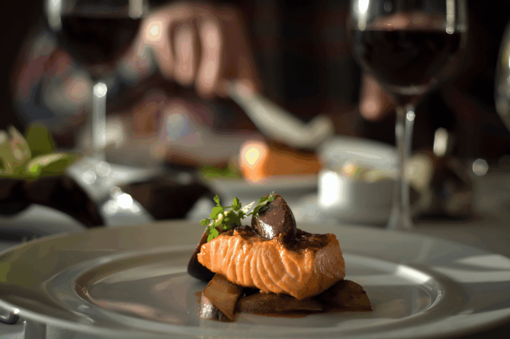 Close-up of a gourmet salmon dish garnished with herbs, mushrooms, and vegetables, served on a white plate in a fine dining setting. Red wine glasses and elegant plating evoke a refined yet intimate atmosphere, highlighting the sensory pleasure and quiet luxury that distinguish fine dining from Michelin’s structured precision.